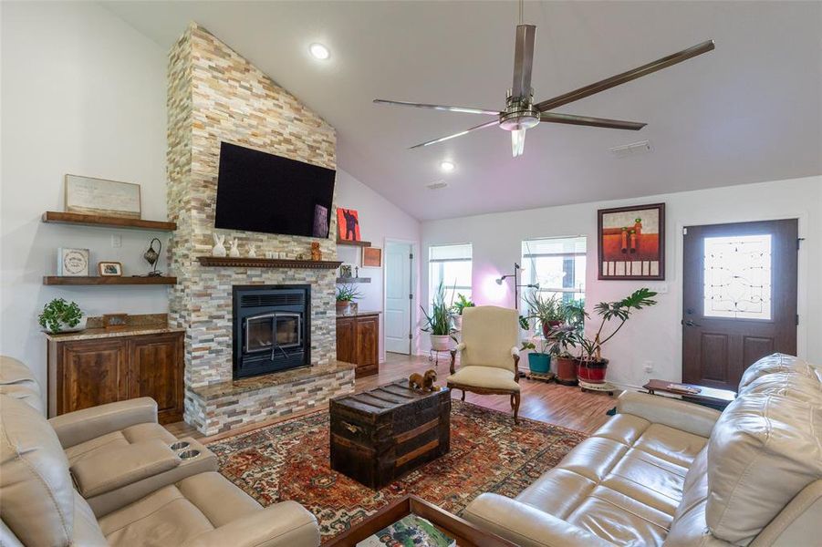 Vaulted ceiling living area featuring a stacked stone fireplace with a raised hearth, wood-finish flooring, and a multi-blade ceiling fan Vaulted ceiling living area featuring a stacked stone fireplace with a raised hearth, wood-finish flooring, and a multi-blade ceiling fan