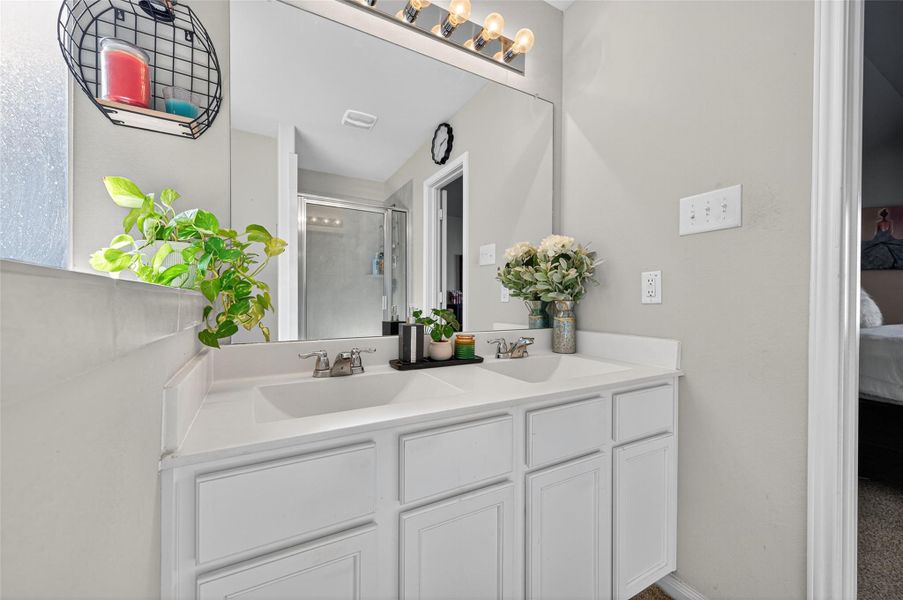 This bathroom features a modern dual-sink vanity with white cabinetry and a large mirror. It includes stylish lighting, a glass-enclosed shower, and decorative touches like plants and candles, creating a fresh and inviting atmosphere.