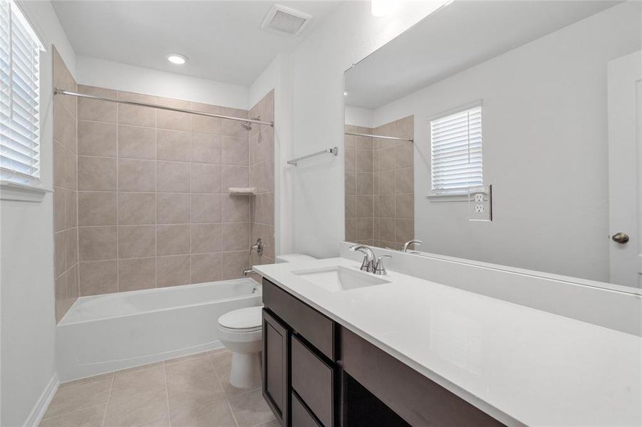 Secondary bath features tile flooring, bath/shower combo with tile surround, stained wood cabinets, beautiful light countertops, mirror, sleek fixtures and modern finishes.