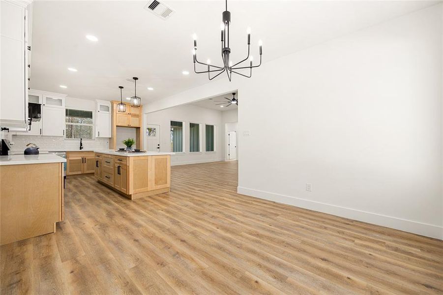 This photo showcases a modern, open-concept living area with light wood flooring, a spacious kitchen featuring a central island with pendant lighting, and large windows for natural light. The space is bright and airy with a contemporary chandelier and fresh white walls.