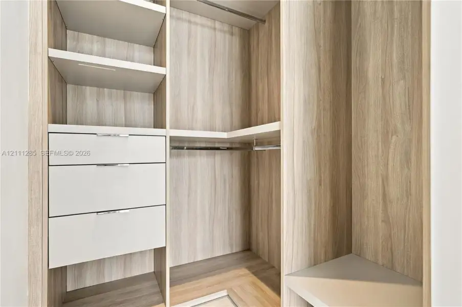 Custom walk-in closet featuring wood-finish cabinetry, open shelving, integrated drawers with metal pulls, and built-in hanging rods Custom walk-in closet featuring wood-finish cabinetry, open shelving, integrated drawers with metal pulls, and built-in hanging rods