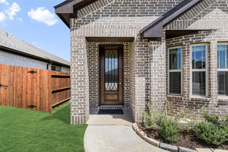 Front entry of brick home with wood and glass door, concrete walkway, and landscaped yard