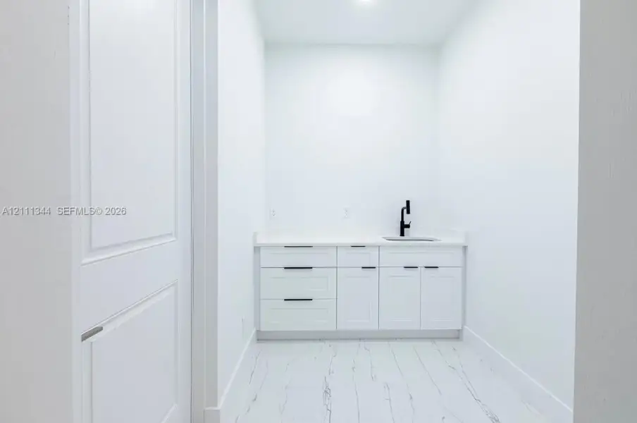 Utility space featuring white shaker-style cabinetry, solid-surface countertops, an undermount sink with a matte black faucet, and large-format tile flooring Utility space featuring white shaker-style cabinetry, solid-surface countertops, an undermount sink with a matte black faucet, and large-format tile flooring