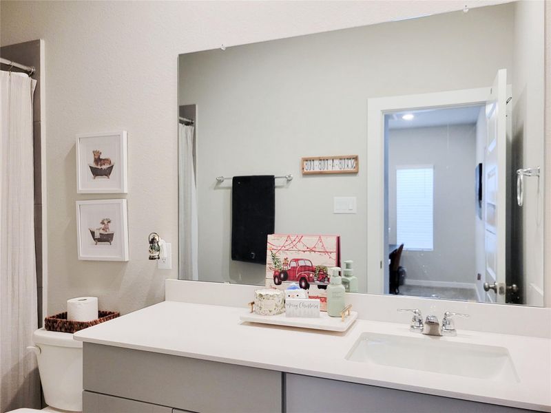 This bathroom features a modern design with a large mirror, sleek white countertop, and neutral wall color. It includes decorative touches like framed art, a stylish tray with essentials, and a cozy atmosphere. The room is well-lit and offers a glimpse into an adjacent space.