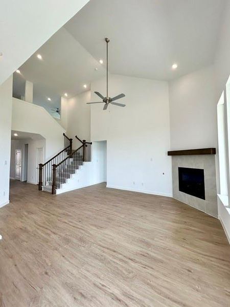 Spacious living area featuring high vaulted ceilings with recessed lighting, wood-finish flooring, a contemporary fireplace with a tile surround and wood mantel, a ceiling fan, and a grand staircase with dark wood railings and balusters Spacious living area featuring high vaulted ceilings with recessed lighting, wood-finish flooring, a contemporary fireplace with a tile surround and wood mantel, a ceiling fan, and a grand staircase with dark wood railings and balusters