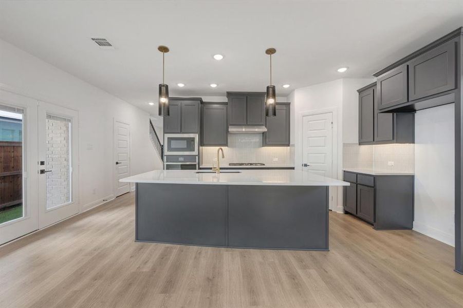 Modern kitchen featuring a large central island with a white countertop, dark gray cabinetry, stainless steel appliances, and wood-finish flooring