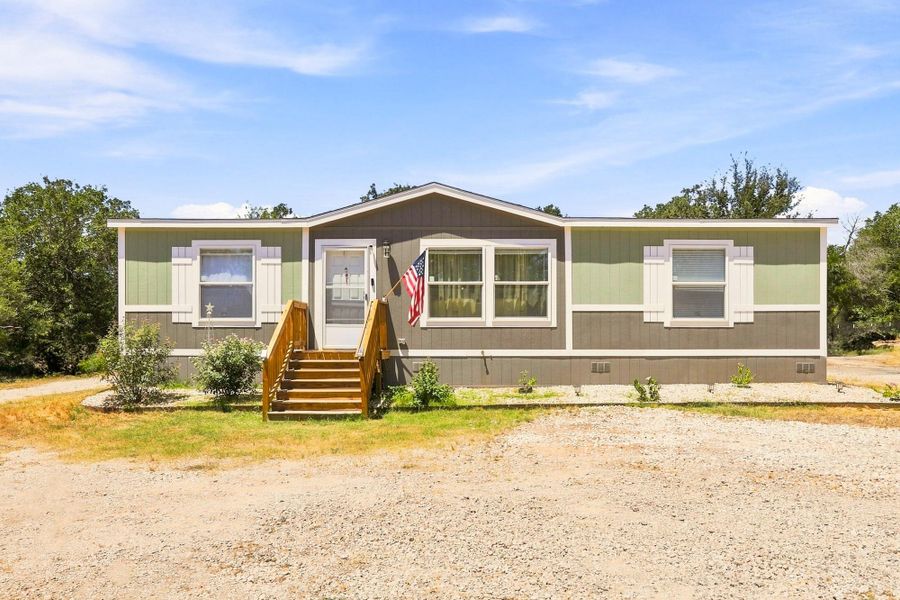 A manufactured home with vertical siding, front steps, and a gravel driveway under clear skies.