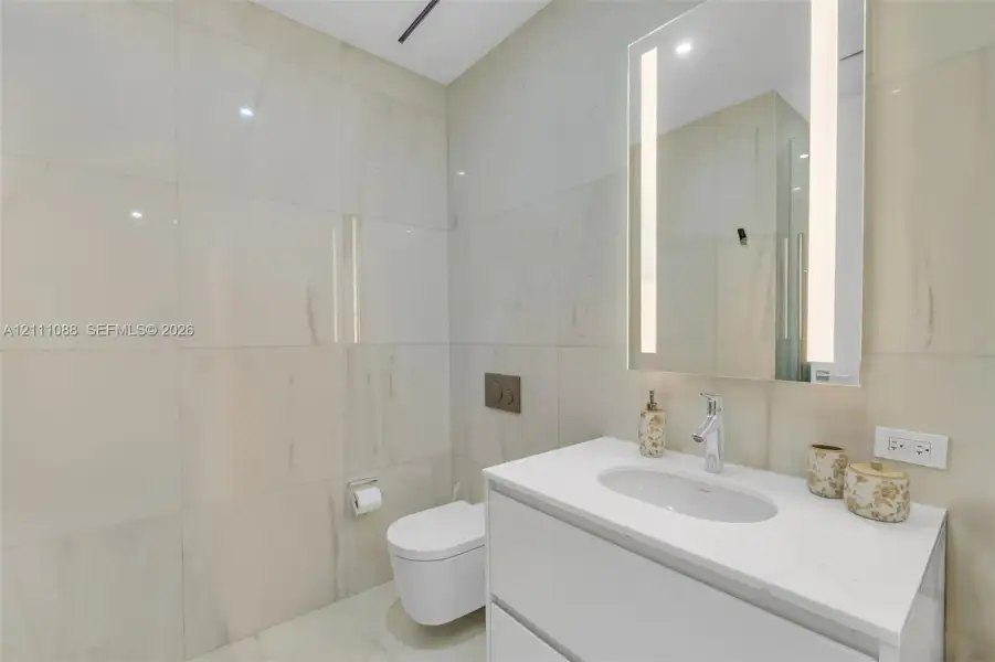 Full-height wall tiling, wall-mounted commode with a dual-flush actuator plate, floating vanity with a white countertop and integrated sink, and an LED-integrated vanity mirror Full-height wall tiling, wall-mounted commode with a dual-flush actuator plate, floating vanity with a white countertop and integrated sink, and an LED-integrated vanity mirror