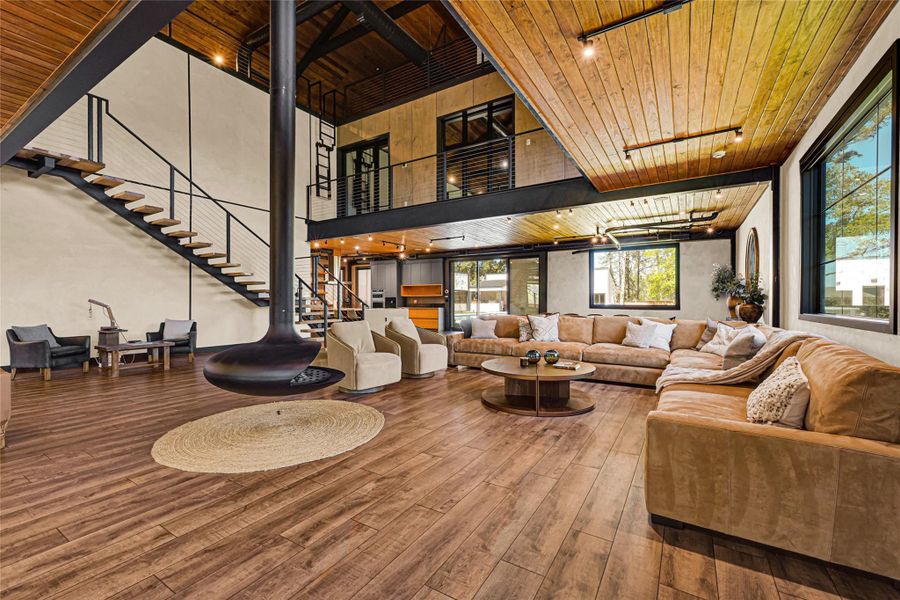 Living area features an open floor plan with high ceilings, a modern suspended fireplace, and a stylish floating staircase. The room is accented with a large sectional sofa, hardwood floors, and abundant natural light from large windows, creating a warm and inviting atmosphere perfect for entertaining.