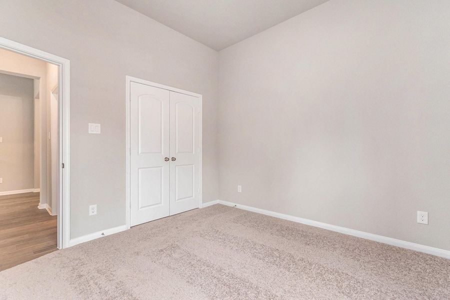 This room features soft beige walls and carpet flooring, offering a cozy atmosphere. It includes a double-door closet and an entryway leading to a hallway with hardwood flooring.