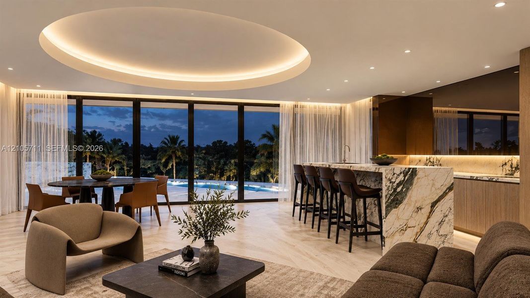Expansive interior featuring a recessed circular tray ceiling with integrated perimeter lighting, floor-to-ceiling glass wall, light-toned wood-finish flooring, and a large kitchen island with a stone-surfaced waterfall edge