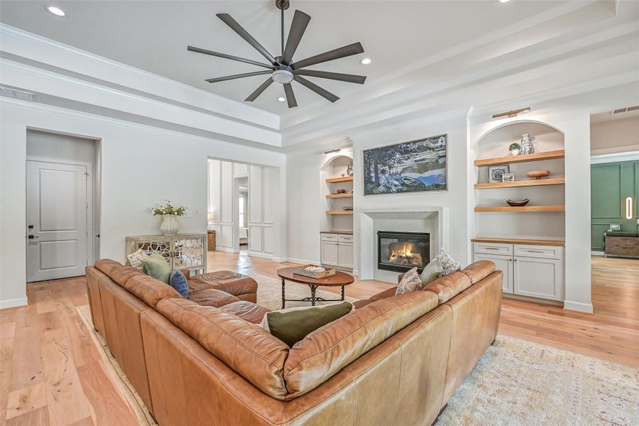 The spacious family room centers around a marble-look gas-log fireplace flanked by custom built-ins with cabinetry, floating wood shelves & accent lighting, while a dramatic tray ceiling and oversized fan add architectural interest overhead.