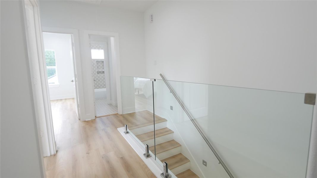 This photo shows a bright, modern hallway with light wood flooring and a glass-paneled staircase. It leads to a bedroom and a bathroom with contemporary tile work, providing a clean and open feel. This photo shows a bright, modern hallway with light wood flooring and a glass-paneled staircase. It leads to a bedroom and a bathroom with contemporary tile work, providing a clean and open feel.