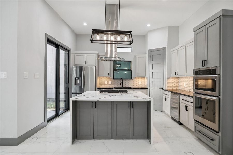 Natural light from the sliding doors enhances the clean lines of the cabinetry and the striking waterfall island. Stainless steel appliances and double ovens offer everyday convenience, while the vent hood and brick-pattern backsplash add character.