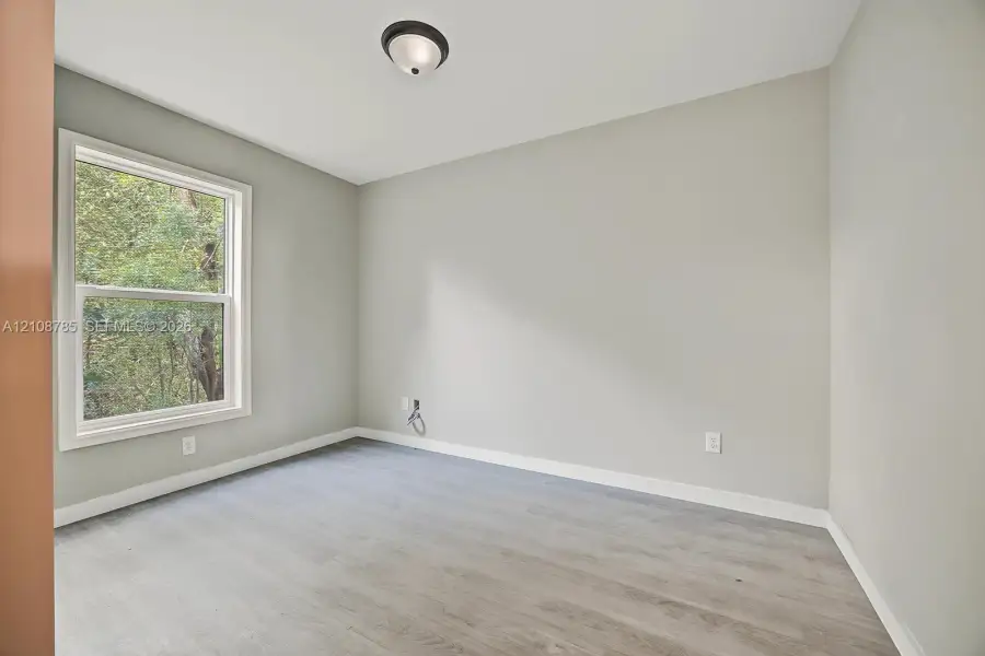Neutral-toned interior space featuring wood-finish flooring, a single-hung window with exterior greenery, and a flush-mount ceiling light fixture Neutral-toned interior space featuring wood-finish flooring, a single-hung window with exterior greenery, and a flush-mount ceiling light fixture