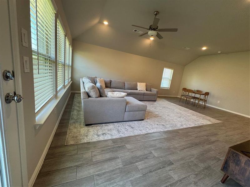 Open-concept living space with wood-finish tile flooring, vaulted ceilings, recessed lighting, and multiple windows with blinds Open-concept living space with wood-finish tile flooring, vaulted ceilings, recessed lighting, and multiple windows with blinds