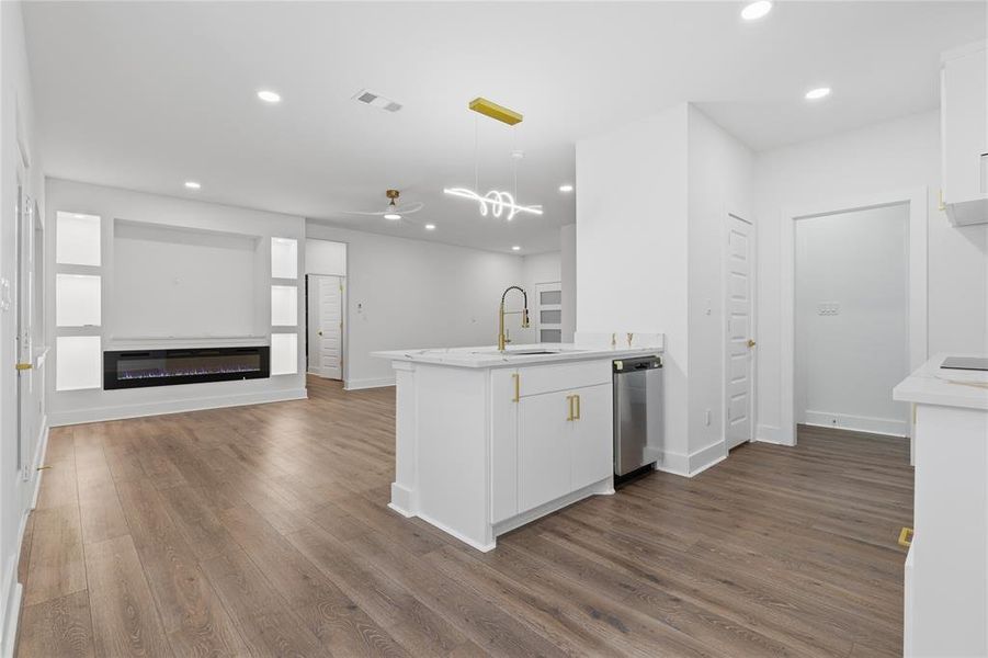 Open-concept living area featuring wood-finish flooring, a contemporary electric fireplace with built-in shelving, a kitchen island with a gold-tone faucet, white cabinetry with gold hardware, and a stainless steel dishwasher