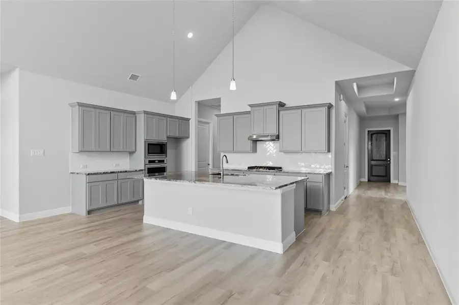 Kitchen featuring gray cabinetry, backsplash, light stone counters, an island with sink, and lofted ceiling