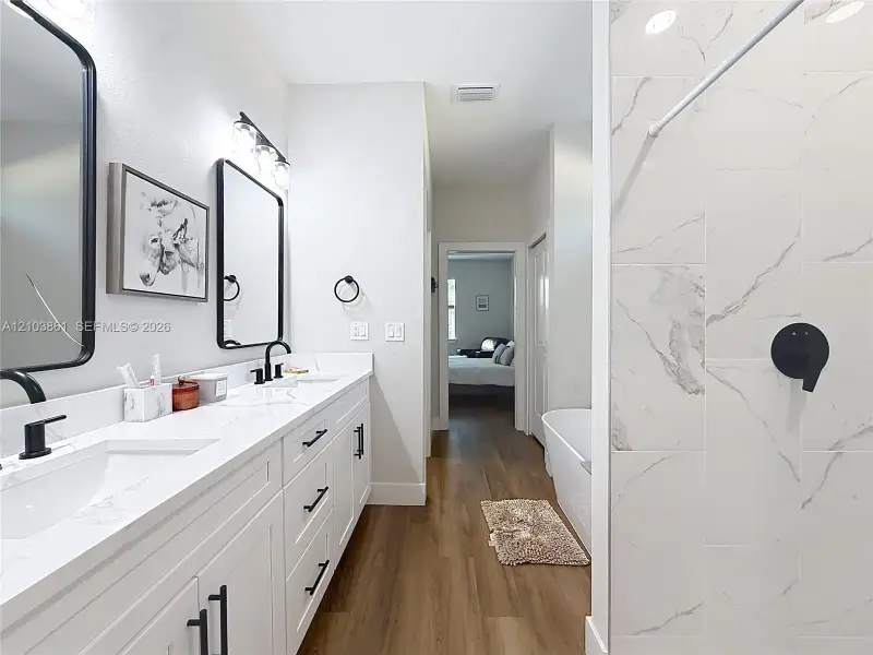 Dual vanity with white cabinetry, stone-finish countertops, matte black fixtures, and framed mirrors, complemented by wood-finish flooring and a tiled shower surround