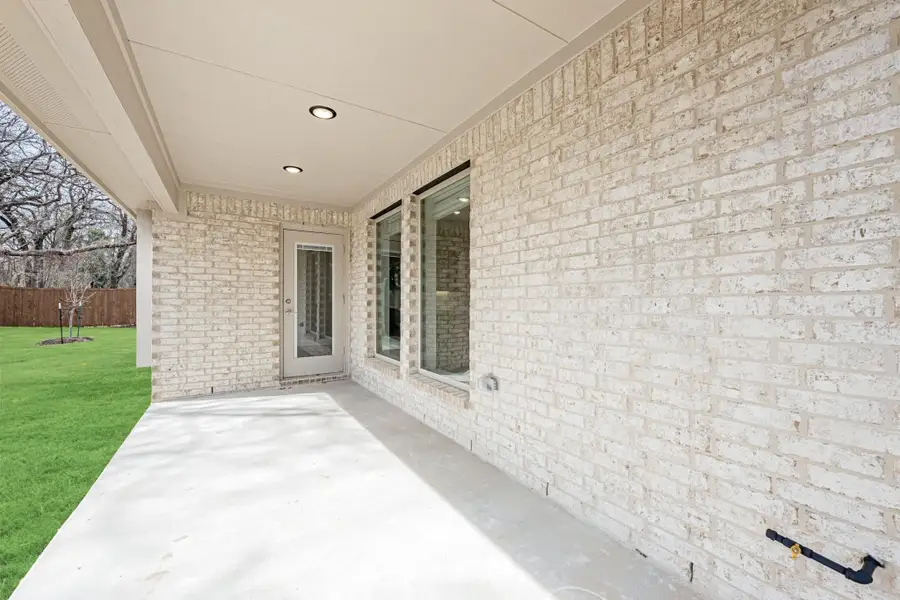 Covered back patio with concrete floor, white brick exterior, recessed lighting, and a glass door overlooking a grassy backyard.
