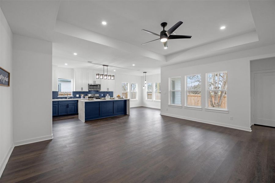 Spacious, open-concept family area features a modern kitchen with bold blue cabinetry, a large island, and sleek lighting. The room is bright with ample natural light from multiple windows, complemented by dark wood flooring and a ceiling fan for comfort. Spacious, open-concept family area features a modern kitchen with bold blue cabinetry, a large island, and sleek lighting. The room is bright with ample natural light from multiple windows, complemented by dark wood flooring and a ceiling fan for comfort.