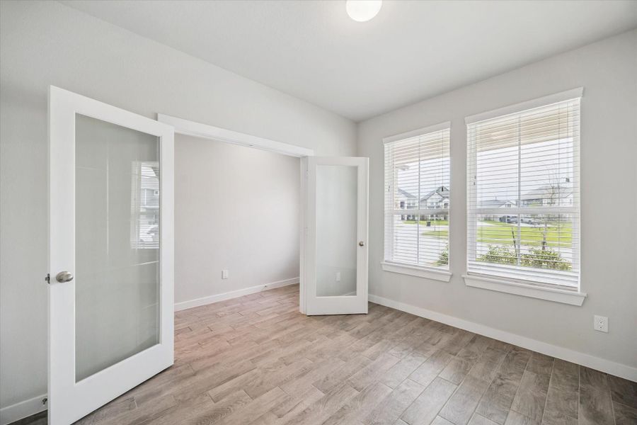 This photo shows a room with wood-style flooring, light gray walls, and two large windows providing natural light. It features glass double doors, making it an ideal space for an office or study. This photo shows a room with wood-style flooring, light gray walls, and two large windows providing natural light. It features glass double doors, making it an ideal space for an office or study.