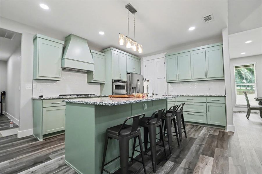 Spacious kitchen featuring an expansive center island, light green cabinetry, stainless steel appliances, a tile backsplash, and wood-finish flooring