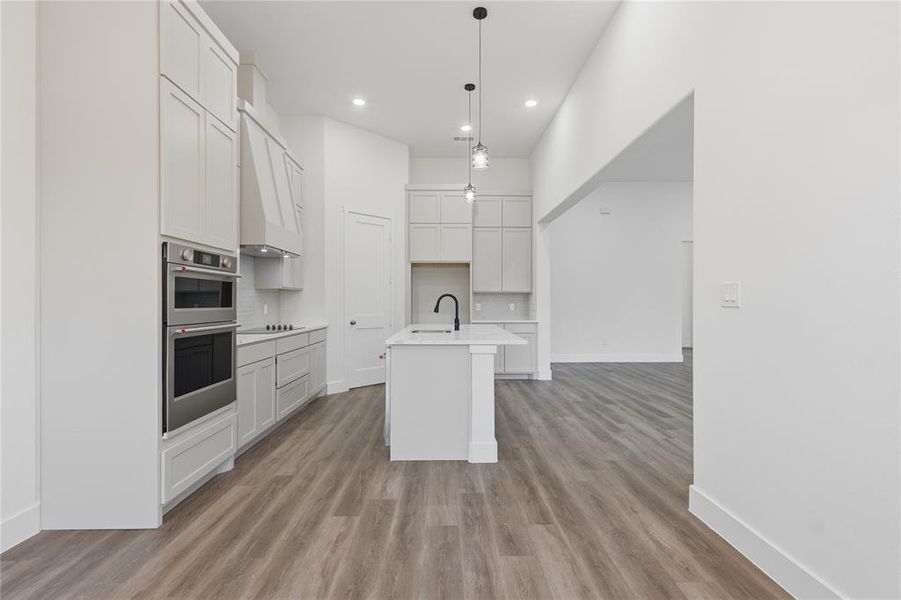 Open-concept kitchen featuring light-toned cabinetry, stainless steel double wall ovens, a center island with a black gooseneck faucet, and wood-finish flooring