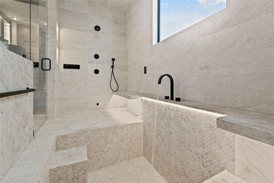 Spa-inspired bathroom featuring large format stone-finish tile walls, a walk-in shower with clear glass enclosure and matte black fixtures, an integrated soaking tub with a matte black deck-mounted faucet, and built-in LED accent lighting