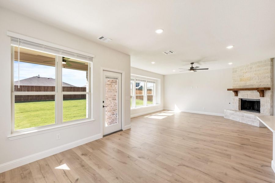 Open living room with light wood floors, stone fireplace, ceiling fan, and large windows overlooking backyard