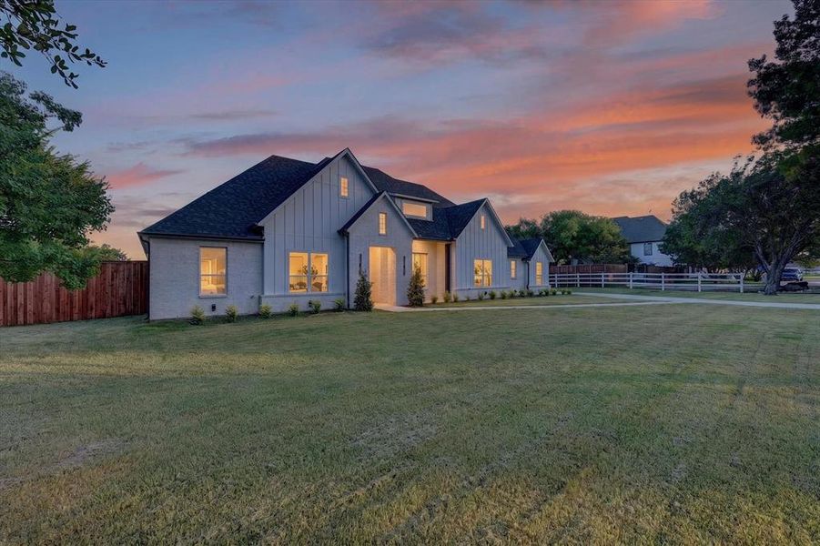 Contemporary farmhouse exterior featuring light brick and vertical board siding, a dark shingle roof, multiple gables, and illuminated windows Contemporary farmhouse exterior featuring light brick and vertical board siding, a dark shingle roof, multiple gables, and illuminated windows