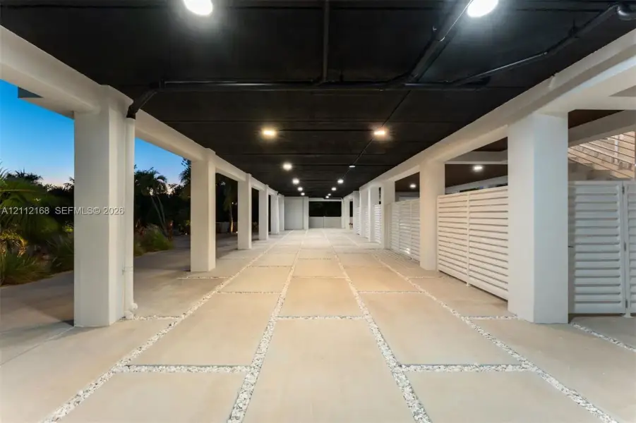 Covered parking area featuring concrete pavers with aggregate inlay, white structural columns, horizontal privacy slat fencing, and exposed dark-finished ceiling Covered parking area featuring concrete pavers with aggregate inlay, white structural columns, horizontal privacy slat fencing, and exposed dark-finished ceiling