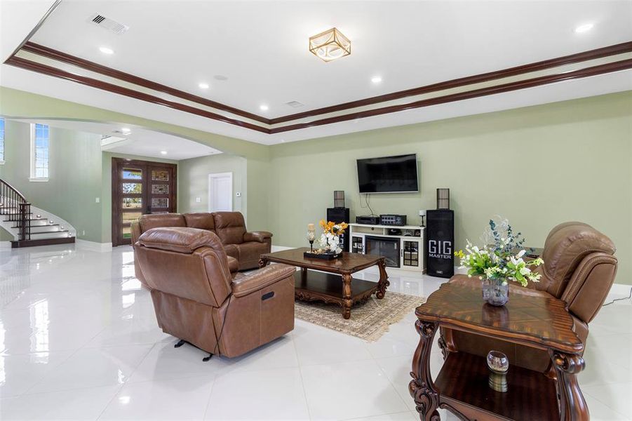 This spacious living room features elegant leather seating, a sleek entertainment setup, and glossy tile floors. It has a stylish coffered ceiling and is well-lit with recessed lighting. The room flows into an open staircase and entryway, creating an inviting and modern atmosphere.