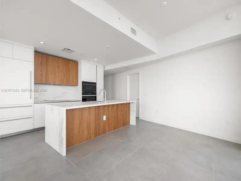 Kitchen featuring a central island with a solid surface waterfall countertop, two-tone cabinetry in white and wood-finish, stone-patterned backsplash, and large-format tile flooring Kitchen featuring a central island with a solid surface waterfall countertop, two-tone cabinetry in white and wood-finish, stone-patterned backsplash, and large-format tile flooring