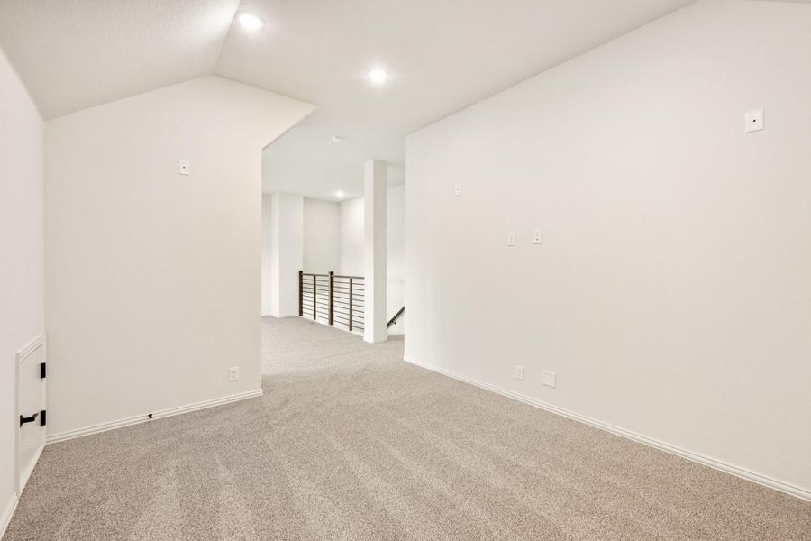 Empty upper-floor loft with vaulted ceiling, gray carpet, and metal stair railing visible in hallway Empty upper-floor loft with vaulted ceiling, gray carpet, and metal stair railing visible in hallway