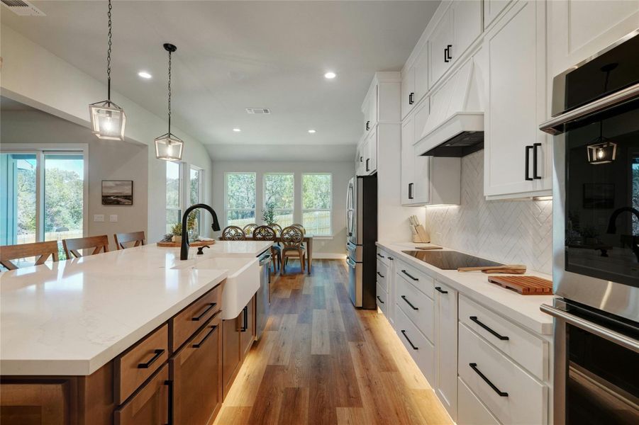 Contemporary kitchen featuring a large island with white countertop and farmhouse sink, wood-finish flooring, white cabinetry with black hardware, and an integrated oven