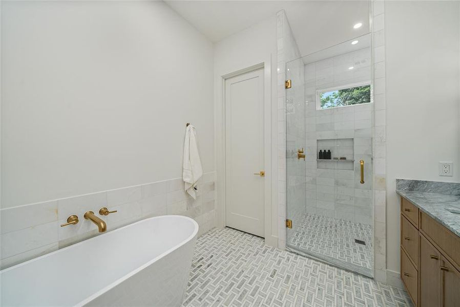 This bathroom features a freestanding bathtub with wall-mounted brass fixtures, a glass-enclosed shower with mosaic tile flooring and a built-in niche, and a vanity with a granite countertop
