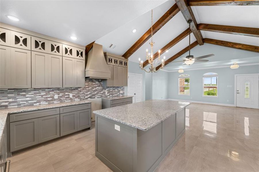 Kitchen featuring vaulted ceiling with beams, backsplash, premium range hood, ceiling fan with notable chandelier, and a center island