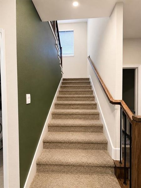 Carpeted staircase featuring a wood-finish handrail with black iron balusters, a window with blinds, and a recessed ceiling light