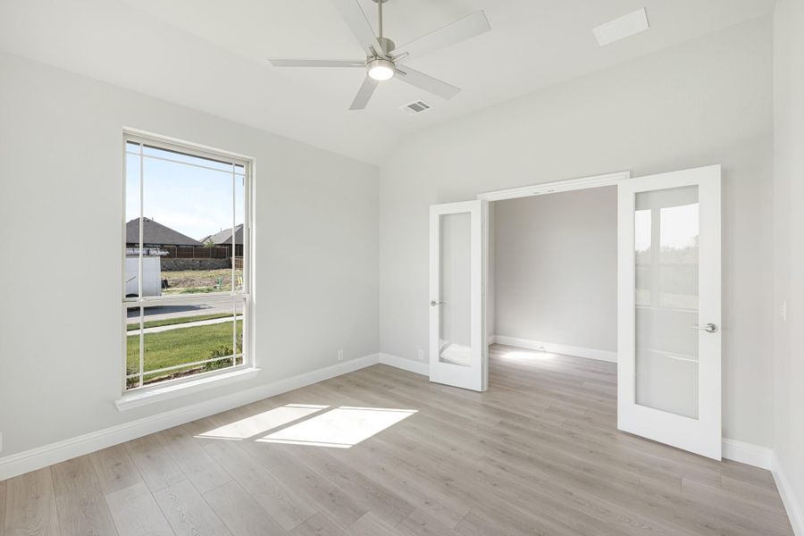 Empty bedroom with light wood floors, ceiling fan, large window, and French doors leading to closet