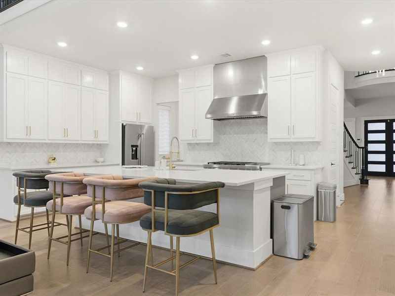 Modern kitchen featuring an expansive center island, white cabinetry, a herringbone tile backsplash, stainless steel appliances, and light wood-finish flooring Modern kitchen featuring an expansive center island, white cabinetry, a herringbone tile backsplash, stainless steel appliances, and light wood-finish flooring
