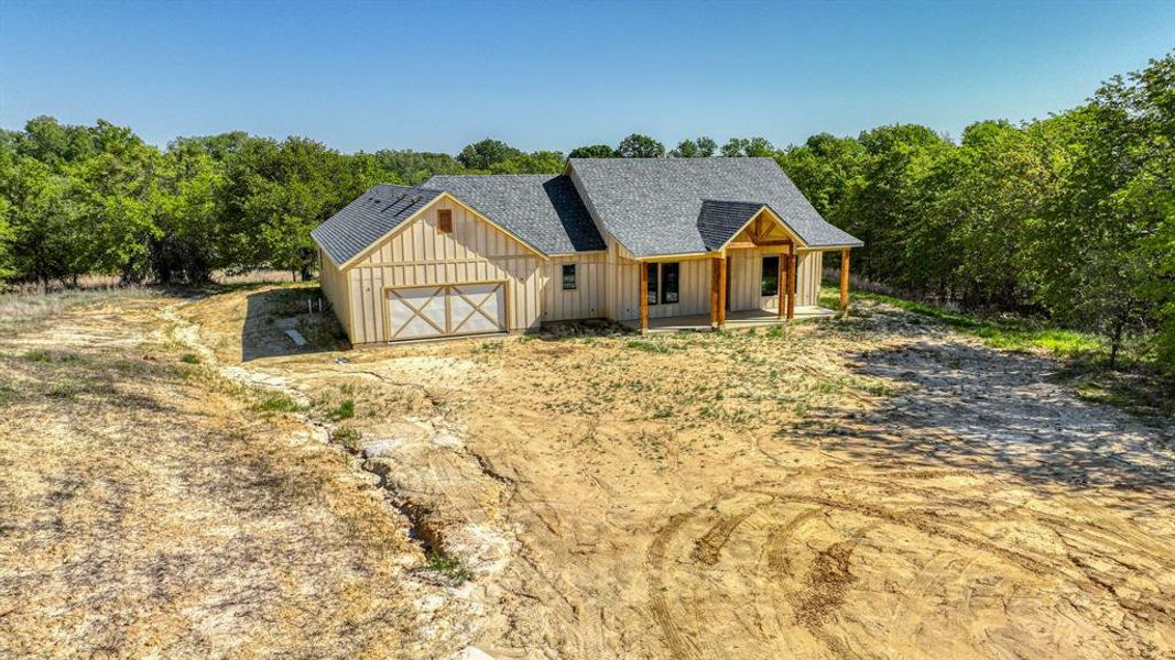 View of front of house with board and batten siding, dirt driveway, roof with shingles, covered porch, and an attached garage View of front of house with board and batten siding, dirt driveway, roof with shingles, covered porch, and an attached garage