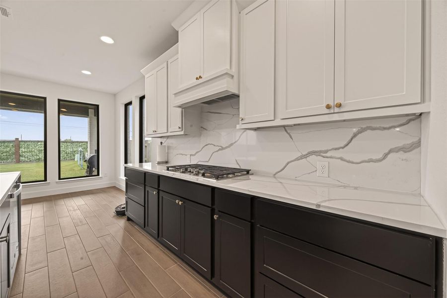 This modern kitchen features sleek Tricon black and Pearl white cabinetry with gold hardware, a marble-patterned backsplash, and a gas stove. Large windows provide ample natural light and a view of the backyard, while wood-look tile flooring adds warmth and style.