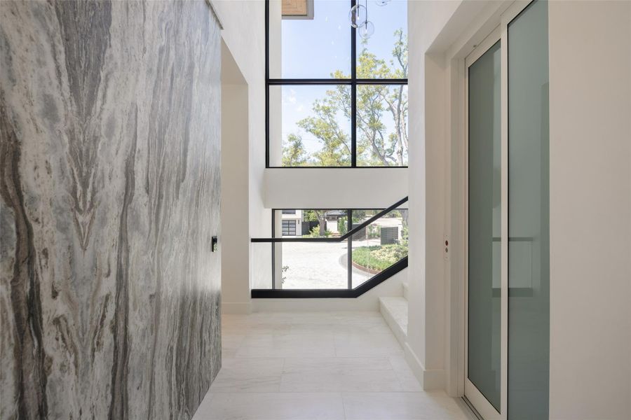 Double-height windows frame tree-lined views beyond the contemporary foyer and stone tile flooring. Double-height windows frame tree-lined views beyond the contemporary foyer and stone tile flooring.