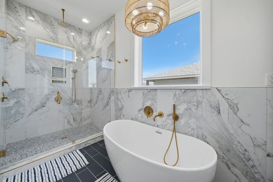 This bathroom features a modern design with elegant marble walls and a spacious glass-enclosed shower. A luxurious freestanding bathtub sits beneath a large window, providing natural light. Warm gold fixtures and a stylish chandelier add a touch of sophistication.