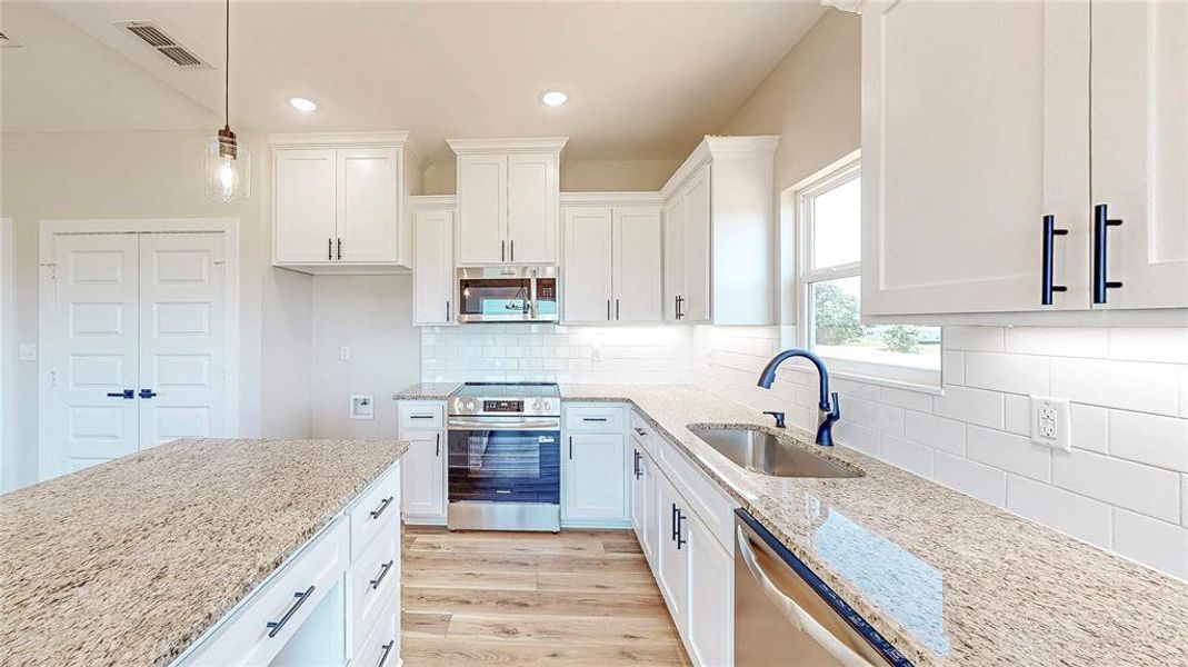 Kitchen with appliances with stainless steel finishes, light wood-type flooring, light stone counters, white cabinetry, and hanging light fixtures Kitchen with appliances with stainless steel finishes, light wood-type flooring, light stone counters, white cabinetry, and hanging light fixtures