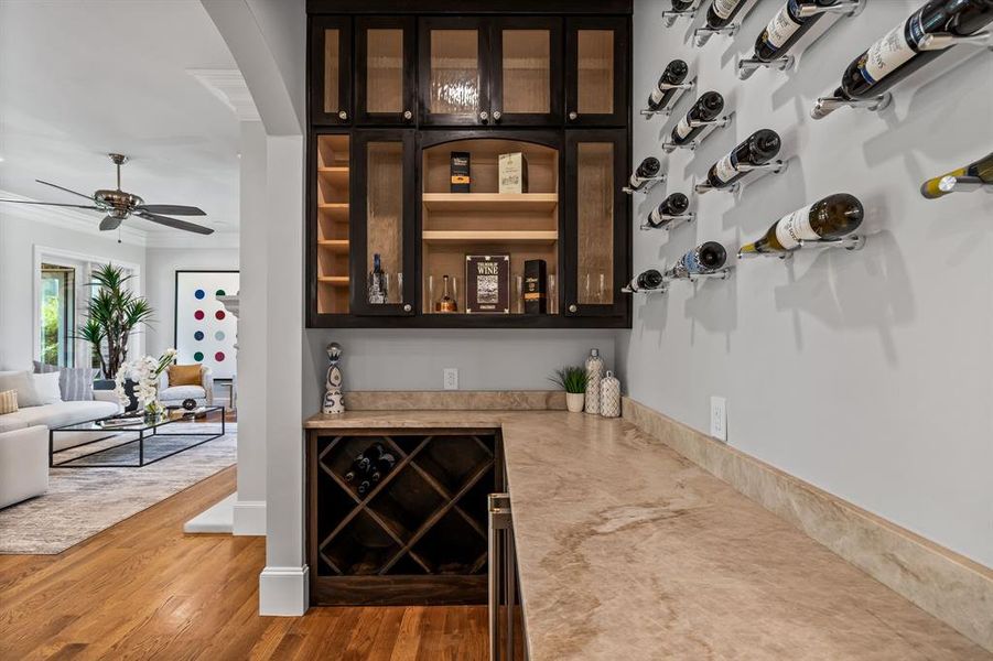 Indoor dry bar featuring glass insert cabinets, open shelves, beverage cooler, wood finished floors, and dark brown cabinetry Indoor dry bar featuring glass insert cabinets, open shelves, beverage cooler, wood finished floors, and dark brown cabinetry