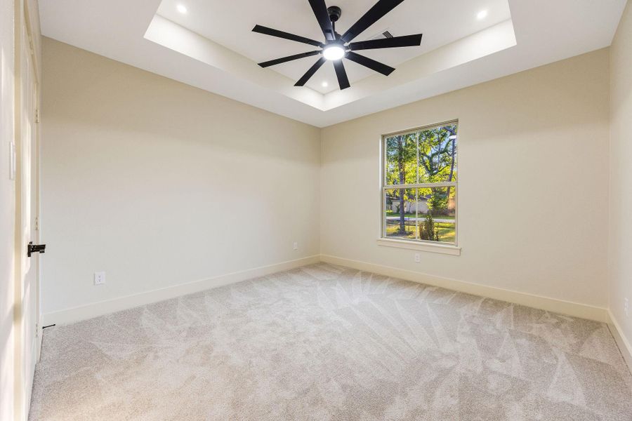 This secondary bedroom features soft carpet flooring, a large window with a view of greenery, and a modern ceiling fan with recessed lighting in a tray ceiling, creating a bright and inviting space.