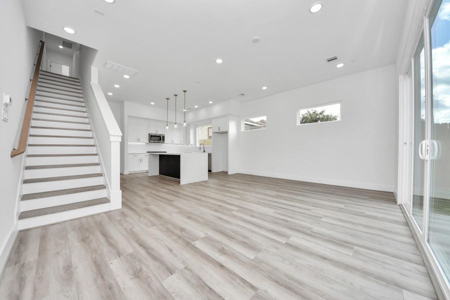 This modern home interior features an open floor plan with light wood flooring, a sleek kitchen with an island, and ample natural light from large windows and sliding glass doors. A staircase with a wooden handrail leads to the upper level.