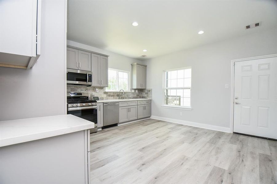 This is a bright, modern kitchen featuring gray shaker cabinets, stainless steel appliances, a subway tile backsplash, and light wood-look flooring. It includes ample storage and counter space, and is complemented by a large window for natural light.